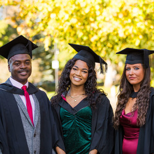 Three graduates standing in the sunshine with their cap and gown on.