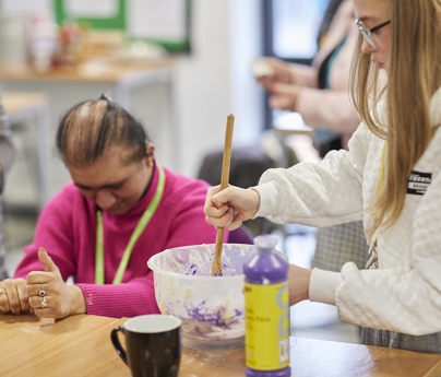 Students in classroom. Some sat down whilst one mixes something in a bowl with purple paint