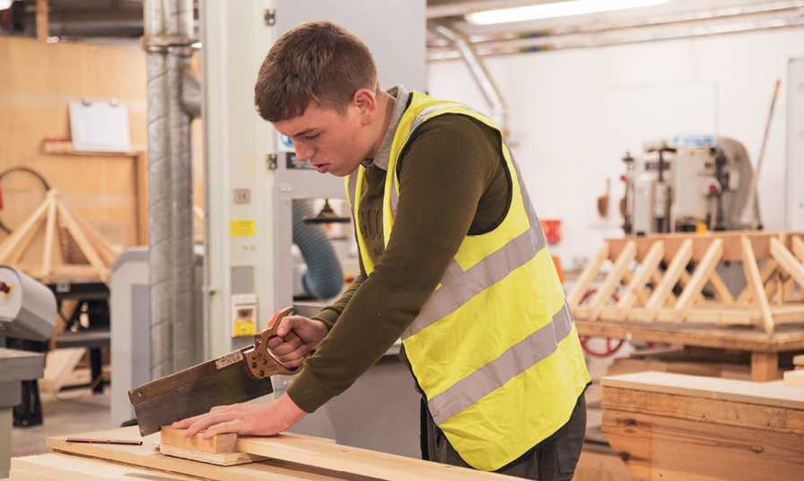 student wearing hi-vis jacket sawing wood in joinery workshop